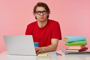 Frowning young student in glasses wears in red t-shirt, man sits by the table and working with laptop, looks at the camera with disgusted, something wrong, isolated over pink background. photo