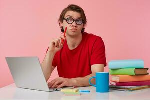 Young thinking student in glasses wears in red t-shirt, man sits by the table and working with laptop, looks up, holds in hand a pencil, trying to solve a hard equation, isolated over pink background. photo