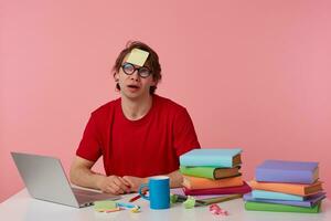 Young thinking man in glasses wears in red t-shirt, sits by the table and working with notebook and books, with a sticker on his forehead, looks up and planing, isolated over pink background. photo