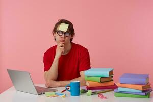 Young thinking man in glasses wears in red t-shirt, with a sticker on his forehead, sits by the table and working with notebook and books, looks up and touches chin, isolated over pink background. photo