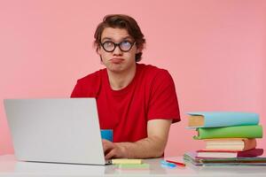 Thinking young student in glasses wears in red t-shirt, man sits by the table and working with laptop, looks up and doubt, isolated over pink background. photo