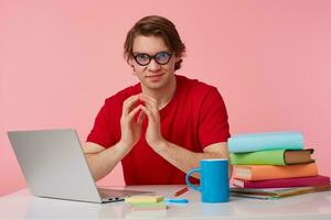 young man in glasses wears in red t-shirt, with a sticker on his forehead, man sits by the table and working with laptop and books, isolated over pink background. Looks displeased and unhappy. photo