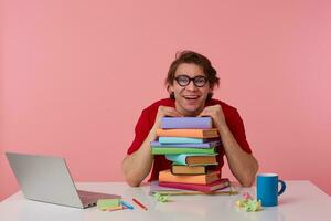 Smiling young man in glasses wears in red t-shirt, man sits by the table and working with laptop and books, leaned on a pile of books, looks happy and glad. Isolated over pink background. photo