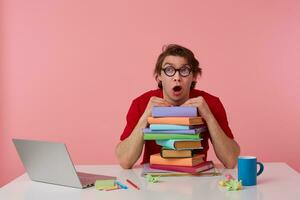 Young guy in glasses, in red t-shirt, sits by the table and working with laptop and books, leaned on a pile of books, looks shocked and surprised with wide open mouth. Isolated over pink background. photo