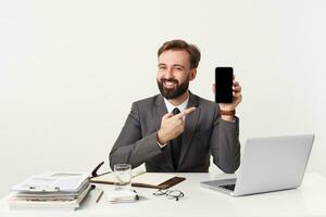 Joyful attractive bearded businessman, top manager sitting at desktop in office, looking camera and smiling, dressed in an expensive suit with a tie, wants to draw your attention to his smartphone. photo