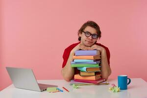 Sad young man in glasses wears in red t-shirt, man sits by the table and working with laptop and books, leaned on a pile of books, isolated over pink background. Looks displeased and unhappy. photo