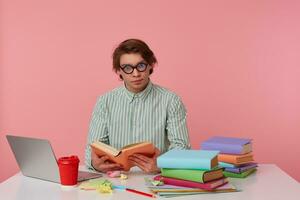 Photo of young man in glasses wears in shirt, student sits by the table and working with notebook, prepared for exam, reads book, having serious look, isolated over pink background.