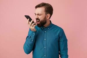 Portrait of an evil young bearded man, wearing a denim shirt, screaming into the phone, messing with someone. Isolated over pink background with copy space . photo
