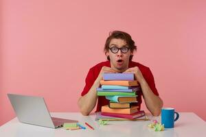 Surprised young guy in glasses, in red t-shirt, man sits by the table and working with laptop and books, leaned on a pile of books, looks shocked with wide open mouth. Isolated over pink background. photo