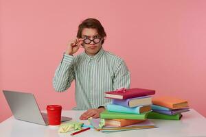 Portrait of disconected man student in glasses wears in red t-shirt, sits by the table and working with notebook and books, prepared for exam, looks through glasses, isolated over pink background. photo