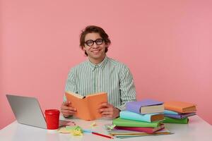 joven sonriente chico en lentes usa en camisa, se sienta por el mesa y trabajando con computadora portátil, preparado para examen, lee libro, mira alegre y disfruta el lectura, aislado terminado rosado antecedentes. foto