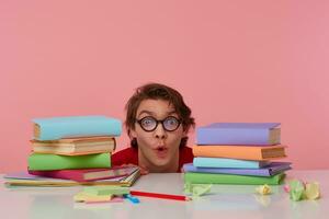 Portrait of happy amazed man in glasses wears in red t-shirt, hiding at the table with books, looks at the camera with surprised expression, looks cheerful, isolated over pink background. photo