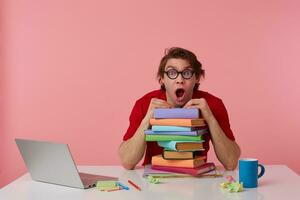 Shocked young man in glasses, in red t-shirt, man sits by the table and working with laptop and books, leaned on a pile of books, looks surprised with wide open mouth. Isolated over pink background. photo