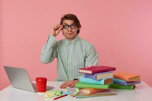 Portrait of smiling man student in glasses wears in red t-shirt, sits by the table and working with notebook and books, prepared for exam, looks through glasses, isolated over pink background. photo