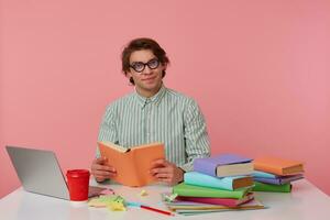 Young smiling man in glasses wears in shirt, student sits by the table and working with notebook, prepared for exam, reads book, looks cheerful and enjoys the reading, isolated over pink background. photo
