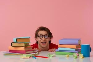 Portrait of positive man in glasses wears in red t-shirt,hiding at the table with books, looks at the camera and smiling, looks cheerful, isolated over pink background. photo