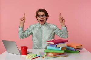 Young shocked student in glasses, sits by the table and working with laptop, looks at the camera and wants draw you attention to copispee over his head, isolated over pink background. photo