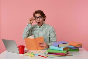 Young wondered student in glasses wears in basic shirt, man sits by the table and working with laptop, looks at the camera through glasses with shocked expression, isolated over pink background. photo