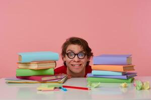 retrato de contento chico en lentes usa en rojo camiseta, ocultación a el mesa con libros, mira a el cámara y sonriente, mira alegre, aislado terminado rosado antecedentes. foto