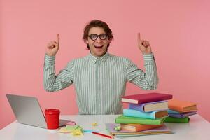 Young happy amazed student in glasses, sits by the table and working with laptop, looks at the camera and wants draw you attention to copispee over his head, isolated over pink background. photo