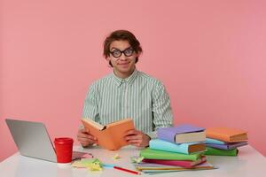 Young smiling man in glasses wears in shirt, student sits by the table and working with notebook, prepared for exam, reads book, having funny look, isolated over pink background. photo