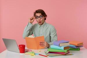 Young shocked student in glasses wears in basic shirt, man sits by the table and working with laptop, looks at the camera through glasses with surpraised expression, isolated over pink background. photo