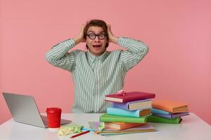 Photo of young shocked guy in glasses, sits by the table and working with laptop, holds his head and looks surprised and scared , isolated over pink background.