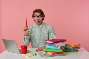 Portrait of young wondersd man in glasses sits by the table and working with laptop, looks at the camera, holds in hand a pencil, have a cool idea, isolated over pink background. photo