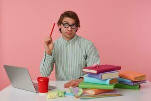 Thinking young man in glasses sits by the table and working with laptop, looks up, holds in hand a pencil, trying to solve a hard equation, isolated over pink background. photo