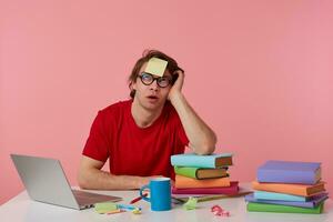 Young pencive man in glasses wears in red t-shirt, sits by the table and working with notebook and books, with a sticker on his forehead, looks up and thinking, isolated over pink background. photo