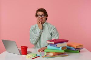 Photo of young afraid guy in glasses, sits by the table and working with laptop, gnawing fingers, looking in camera with scared expression, isolated over pink background.