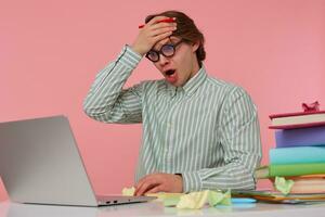 Portrait of man in glasses sits by the table and working with laptop, very dissatisfied, holds hand on forehead with wide open mouth, isolated over pink background. photo