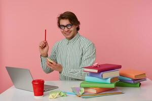 Guy in glasses sits by the table and working with laptop, holds in hand a pencil, have a great idea, looks at the camera and broadly smiling, isolated over pink background. photo