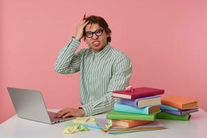 Portrait of angry young guy in glasses sits by the table and working with laptop, very dissatisfied, holds hand on forehead with wide open mouth, isolated over pink background. photo
