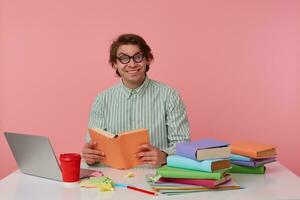 Young cheerful man in glasses wears in shirt, sits by the table and working with notebook, prepared for exam, reads book, looks happy and enjoys the reading, isolated over pink background. photo