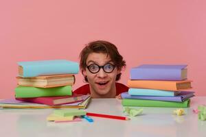 Photo of happy amazed guy in glasses wears in red t-shirt, hiding at the table with books, looks at the camera with wide open mouth, looks cheerful, isolated over pink background.