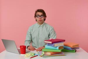 Photo of young sad guy in glasses, sits by the table and working with laptop, looking in camera with unhappy expression, isolated over pink background.