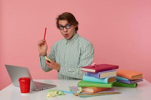 Guy in glasses sits by the table and working with laptop, holds in hand a pencil, have a great idea, looks at the camera, isolated over pink background. photo