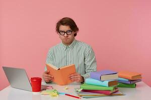 Young doubting man in glasses wears in shirt, sits by the table and working with notebook, prepared for exam, reads book, looks sad and tired, isolated over pink background. photo