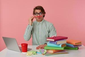 Posirive man in glasses sits by the table and working with laptop, looks at the camera, holds ipencil in lips, looks funny isolated over pink background. photo