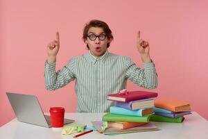 Young surprised student in glasses, sits by the table and working with laptop, looks at the camera and wants draw you attention to copispee over his head, isolated over pink background. photo