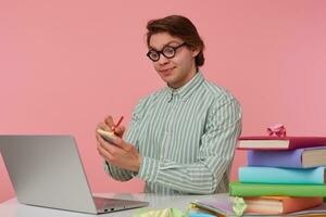 Young glad guy in glasses sits by the table and working with laptop, holds in hand a pencil, have a cool idea, pleased with himself and writes the idea on the sticker, isolated over pink background. photo