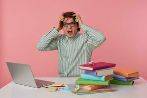 Indoor shot of stressed young dark haired male posing over pink background in striped shirt, sitting at working table with a lot of books, clutching his head with frowned face photo