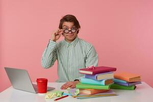 Portrait of positive man student in glasses wears in red t-shirt, sits by the table and working with notebook and books, prepared for exam, looks through glasses, isolated over pink background. photo