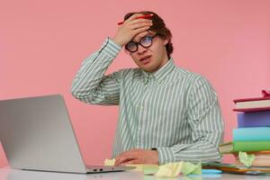 Portrait of displeased young man in glasses sits by the table and working with laptop, very dissatisfied, holds hand on forehead with wide open mouth, isolated over pink background. photo