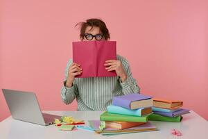 Studio photo of young dark haired male posing over pink background, wearing shirt and glasses, sitting at working table and keeping book in raised hands, looking at camera with wide eyes opened