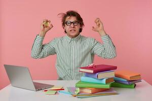 Studio shot of angry young man in eyewear with wild hair sitting at working table, raising hands scowlingly with rumpled notes and looking to camera with grimace, isolated over pink background photo