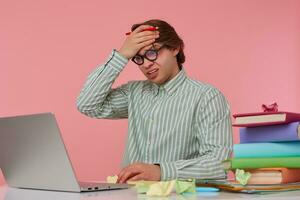 Portrait of man in glasses sits by the table and working with laptop, very dissatisfied with wrongly entered data into the report, holds hand on forehead, isolated over pink background. photo