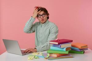 foto de infeliz joven hombre en lentes se sienta por el mesa y trabajando con computadora portátil, muy insatisfecho, sostiene mano en frente aislado terminado rosado antecedentes.