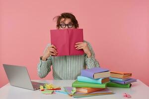 Portrait of young man with dark wild hair reading book over pink background, sitting at working with laptop and preparing for exams, wearing shirt and glasses photo